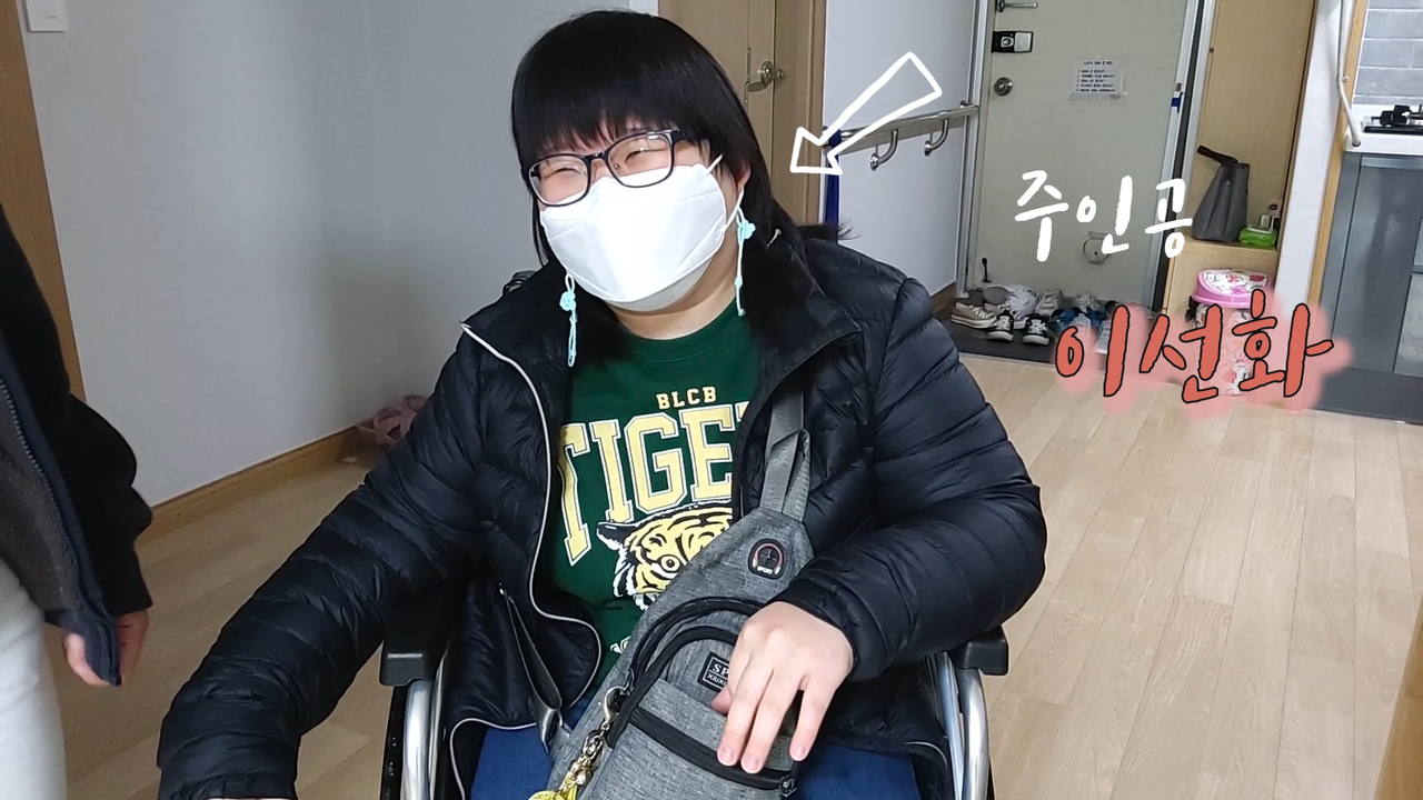휠체어를 탄 사람이 마스크를 쓰고 웃고 있다. 그 사람을 향해 화살표가 그려져 있고 '주인공 이선화'라고 쓰여있다.