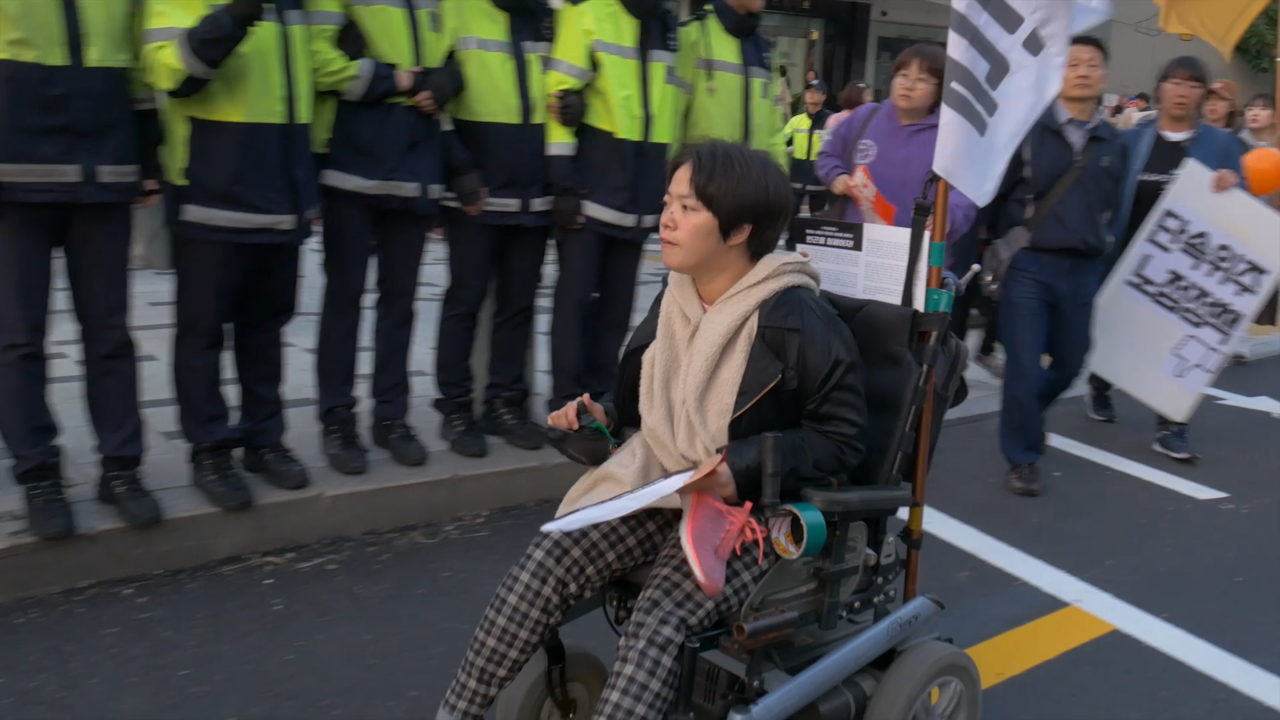 시위 현장의 애린의 모습. 휠체어를 타고 차도를 지나가고 있다. 휠체어 뒤에 깃발이 펄럭인다. 인도에는 경찰들이 몸으로 벽을 만들고 있다.