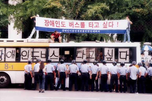 활동가들이 버스 위에서 현수막을 펼치고 있다. 현수막: 장애인도 버스를 타고 싶다! 장애인이동권쟁취를위한연대회의