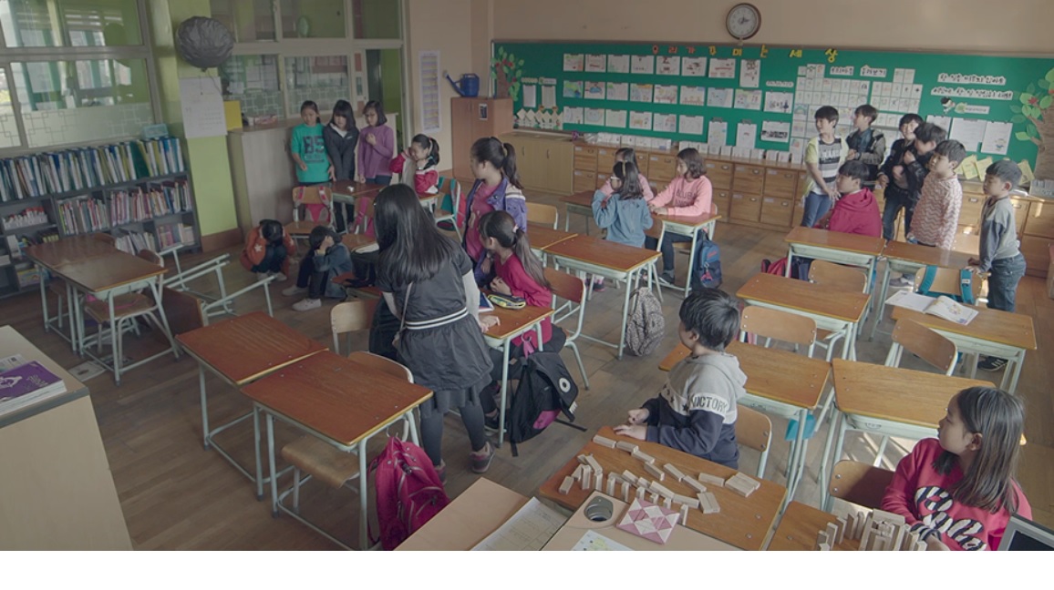 초등학교 교실의 모습. 화면 왼쪽에 책상 두개가 쓰러져 있고 그 뒤에 두 아이가 귀를 막고 주저앉아있다. 다른 학생들이 전부 둘을 쳐다보고 있다.