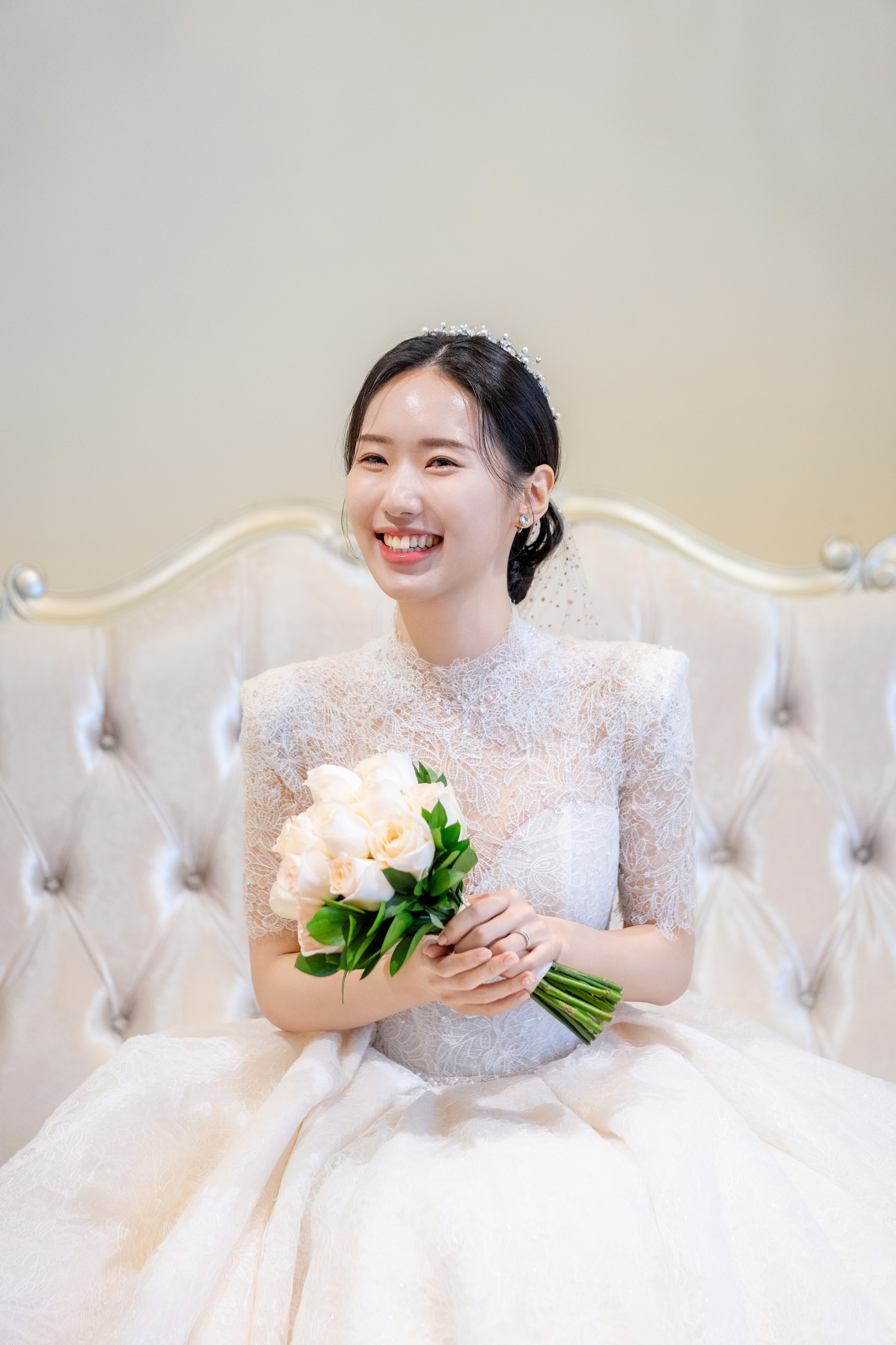 흰 소파에 앉아 부케를 안고 정면을 보며 활짝 웃는 신부