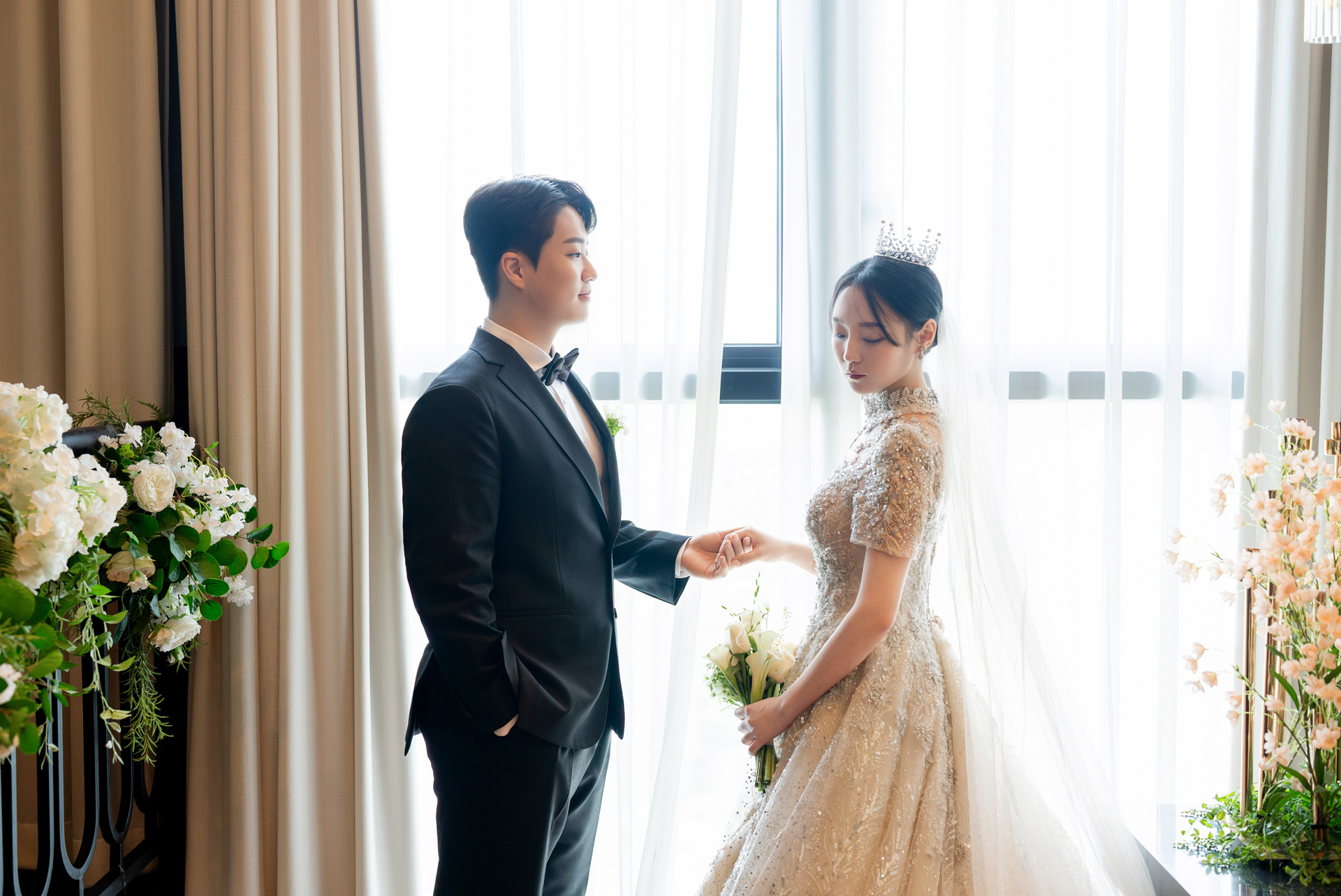 창가 커튼 앞에서 마주 서서 손을 맞잡은 신랑신부