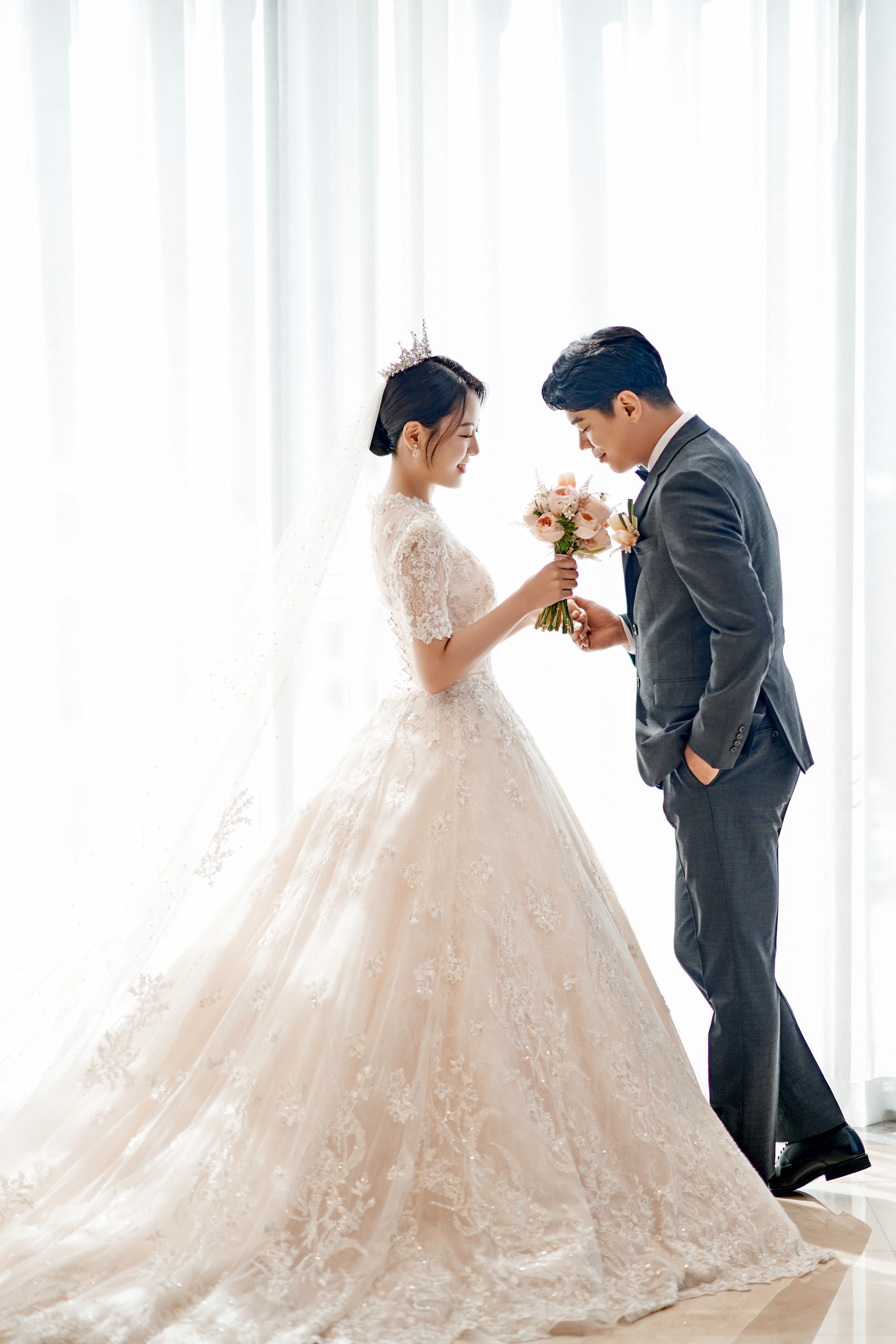 창가 커튼 앞에서 부케를 함께 든 신랑신부, 신부의 트레인이 바닥에 길게 퍼져 있다