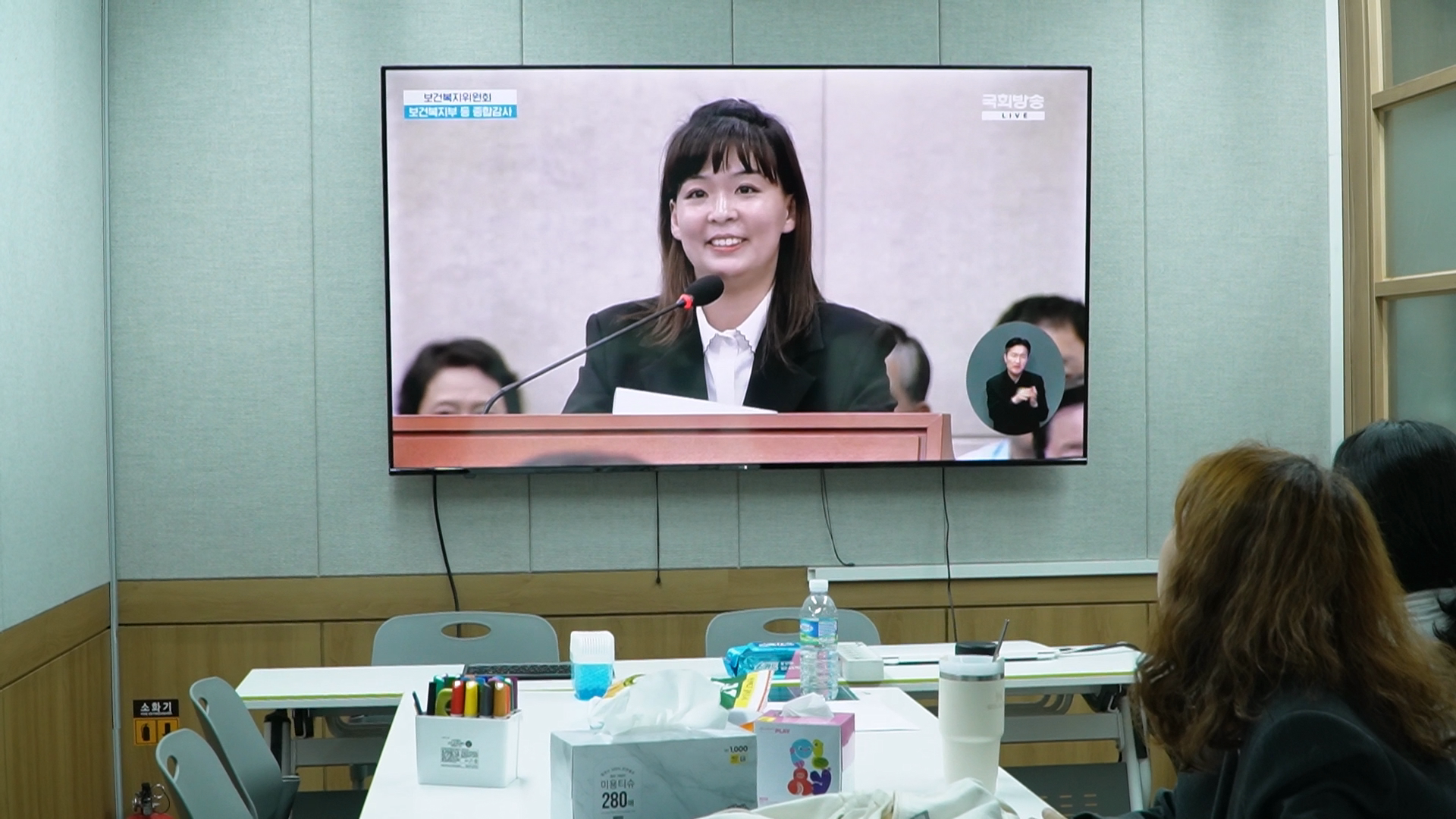 마스크를 쓴 두 사람이 앉아있다. 오른쪽 사람은 녹음용 마이크 앞에 앉아 헤드셋을 끼고 있고, 왼쪽 사람은 어딘가를 가리키고 있다.