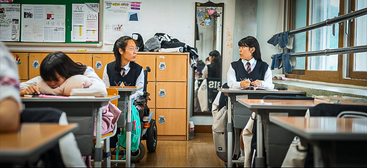 학교 교실. 학생 둘이 서로를 마주보고 있다. 한 학생을 휠체어를 타고 있다.