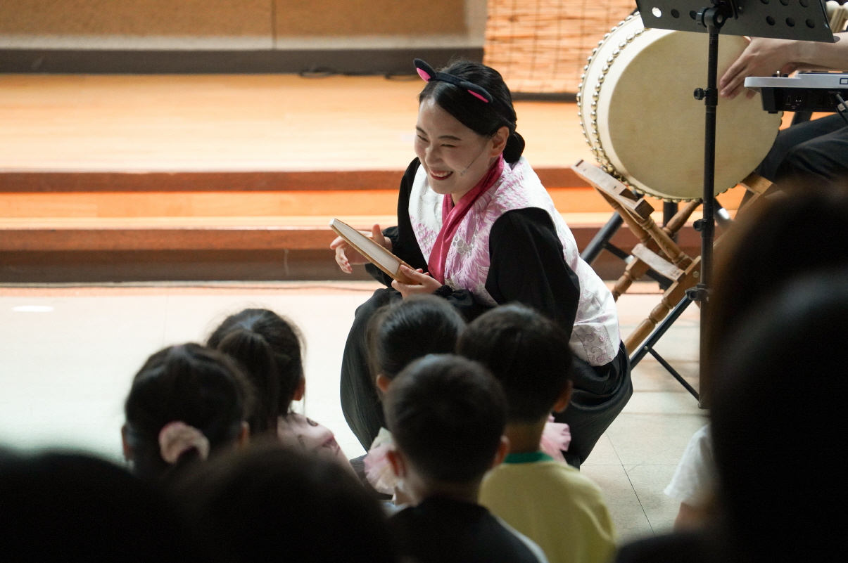 전남 고흥군립중앙도서관 공연 마당놀이 <나는 고양이로소이다> 공연 중 배우가 무대 아래로 내려와 아이들과 눈높이를 맞추며 웃고 있는 모습. 공연과 관객이 가까워진 따뜻한 도서관 문화예술 프로그램의 장면.