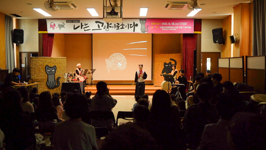 전남 고흥군립중앙도서관 공연 마당놀이 <나는 고양이로소이다> 공연 중, 배우가 무대 중앙에서 고양이 캐릭터와 함께 연기하고 있고 관객들이 집중해 관람하는 모습. 따뜻하고 정겨운 분위기의 도서관 문화공연 현장.