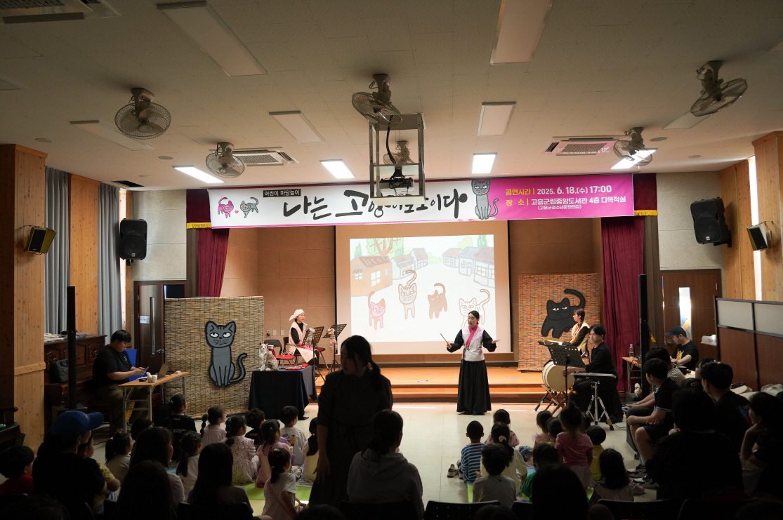 전남 고흥군립중앙도서관 공연 어린이 마당놀이 <나는 고양이로소이다> 현장. 무대 앞에 아이들이 앉아 있고 배우들이 고양이 애니메이션 배경 앞에서 공연 중인 모습. 도서관 강당이 마을 사랑방처럼 따뜻한 분위기를 이루고 있다.