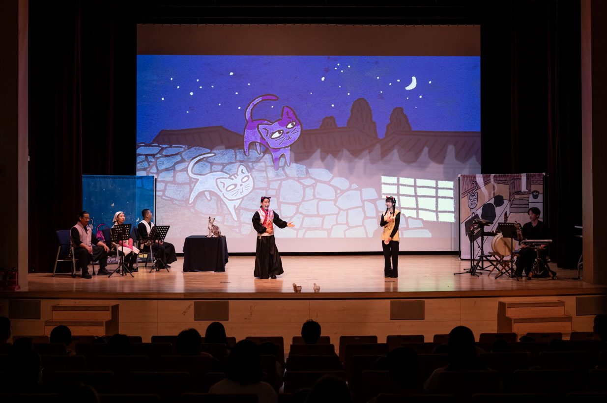 어린이 마당놀이 〈나는 고양이로소이다〉 공연 장면. 밤하늘과 담장을 배경으로 길고양이들이 등장하는 애니메이션이 무대 뒤에 투사되고, 고양이 역할의 배우들이 무대 중앙에서 이야기를 전개하고 있다. 객석에는 가족 단위 관객들이 공연을 관람 중이다.