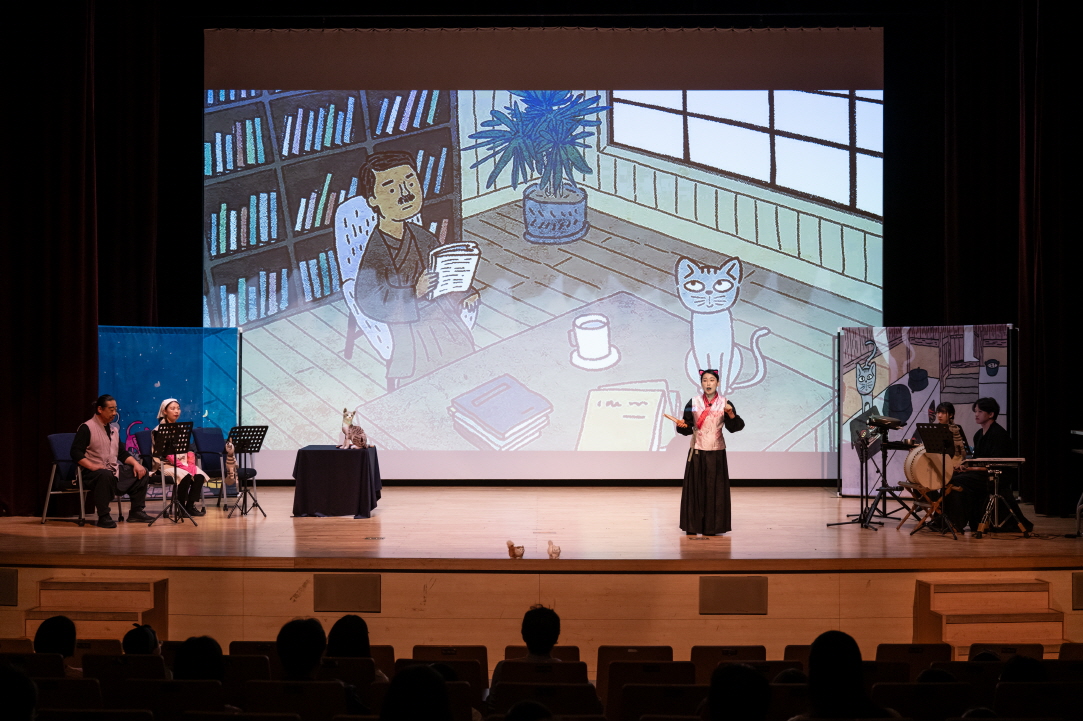 어린이 마당놀이 〈나는 고양이로소이다〉 공연 장면. 고양이 주인공이 새로운 가족인 작가 아저씨를 만나 함께 살아가게 된 이야기를 전개하는 무대. 배경에는 책장과 책상이 있는 서재 애니메이션이 투사되어 있으며, 배우는 관객을 향해 대사를 전달하고 있다.