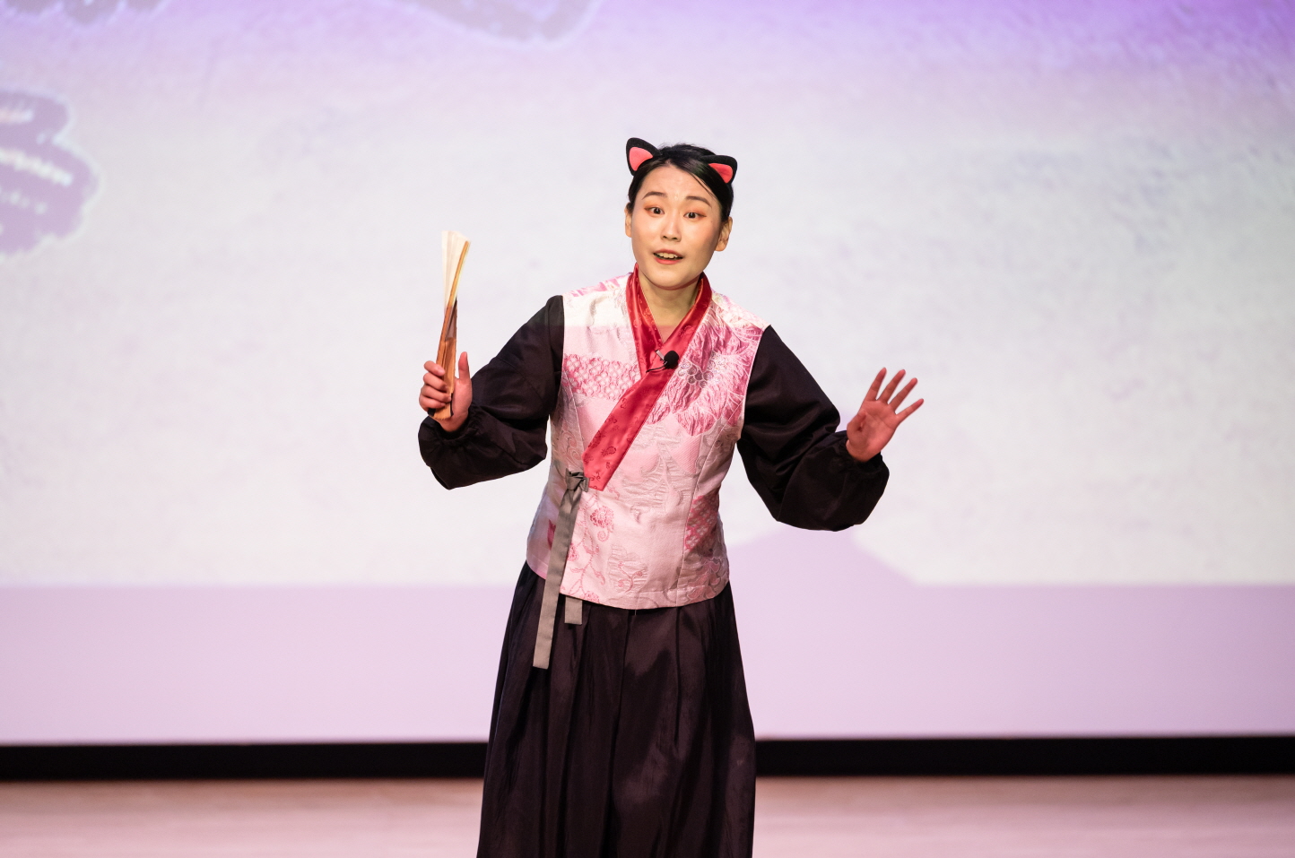 고양이 귀 머리띠를 착용한 배우가 어린이 마당놀이 〈나는 고양이로소이다〉 공연 중 관객을 향해 손짓하며 이야기하고 있는 장면. 손에는 부채를 들고 전통 의상을 입고 무대 위에 서 있다.