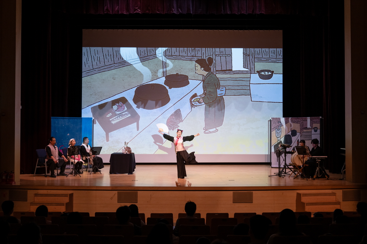 전주 한국전통문화전당에서 열린 어린이 마당놀이 〈나는 고양이로소이다〉 공연 장면. 고양이로 분장한 배우가 전통 부엌 배경의 애니메이션 화면 앞에서 관객과 소통하며 연기하고 있으며, 양옆에는 연주자와 출연진이 함께 무대를 구성하고 있다.
