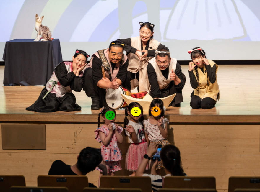 어린이 마당놀이 〈나는 고양이로소이다〉 공연 후 무대에서 고양이 분장의 배우들과 어린이 관객들이 함께 포즈를 취하며 기념사진을 찍는 모습. 배우들은 귀여운 고양이 포즈를 하고 있고, 아이들은 밝은 표정으로 앞줄에 서서 사진 촬영에 응하고 있다.