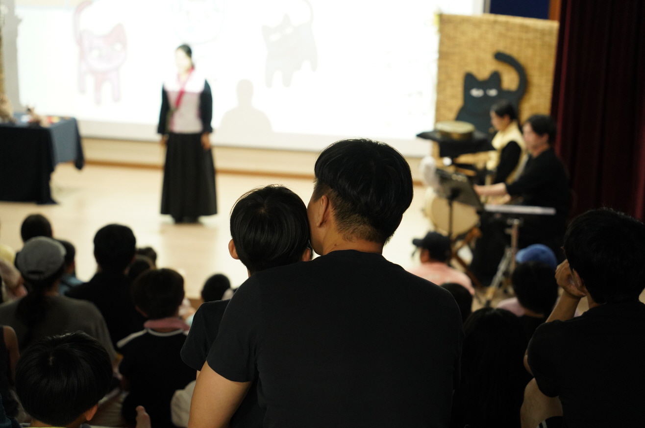순천 기적의도서관에서 나는고양이로소이다. 공연을 관람 중인 아빠와 아이. 고양이 일러스트 배경 앞에서 배우가 공연 중이다."
