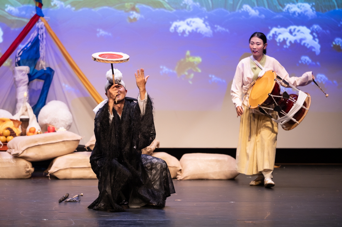 마당놀이 지신지신 출연자가 버나 접시돌기기 묘기를 선보이는 장면, 장구 연주자가 흥겹게 장단을 맞추며 코믹하고 흥겨운 분위기 연출