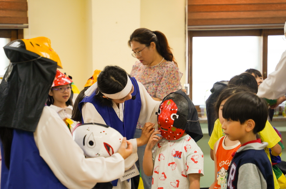 초등학교 K-POP 탈춤체험 – 강사가 저학년 어린이에게 전통 탈을 씌워주는 모습