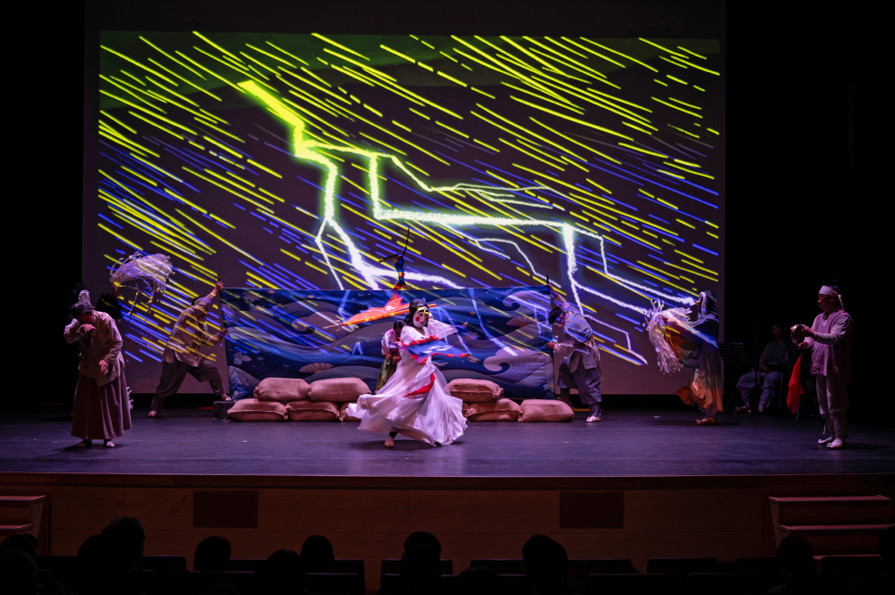 번개와 폭우가 내리는 영상 배경 속에서 흰 탈 캐릭터가 무대 중앙에서 천을 휘날리며 역동적으로 춤추는 마당놀이 지신지신 장면