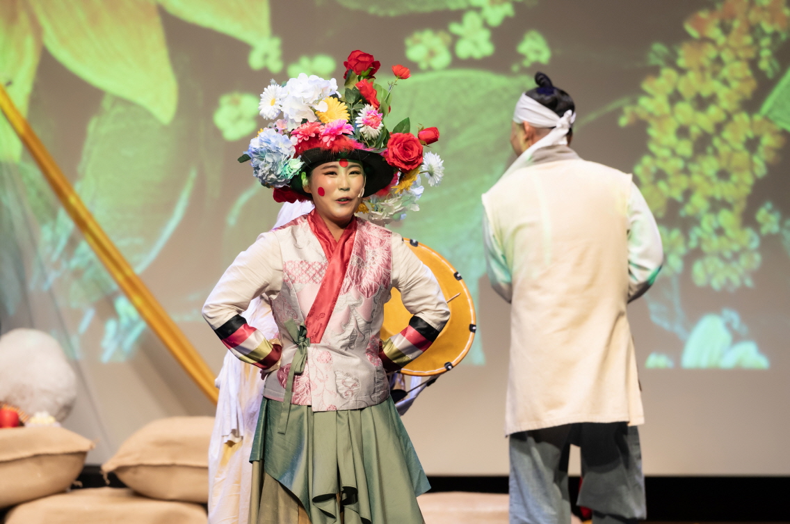 화려한 꽃 장식을 머리에 쓴 배우가 익살스러운 표정과 전통의상을 입고 무대에서 연기하는 마당놀이 지신지신 공연 장면