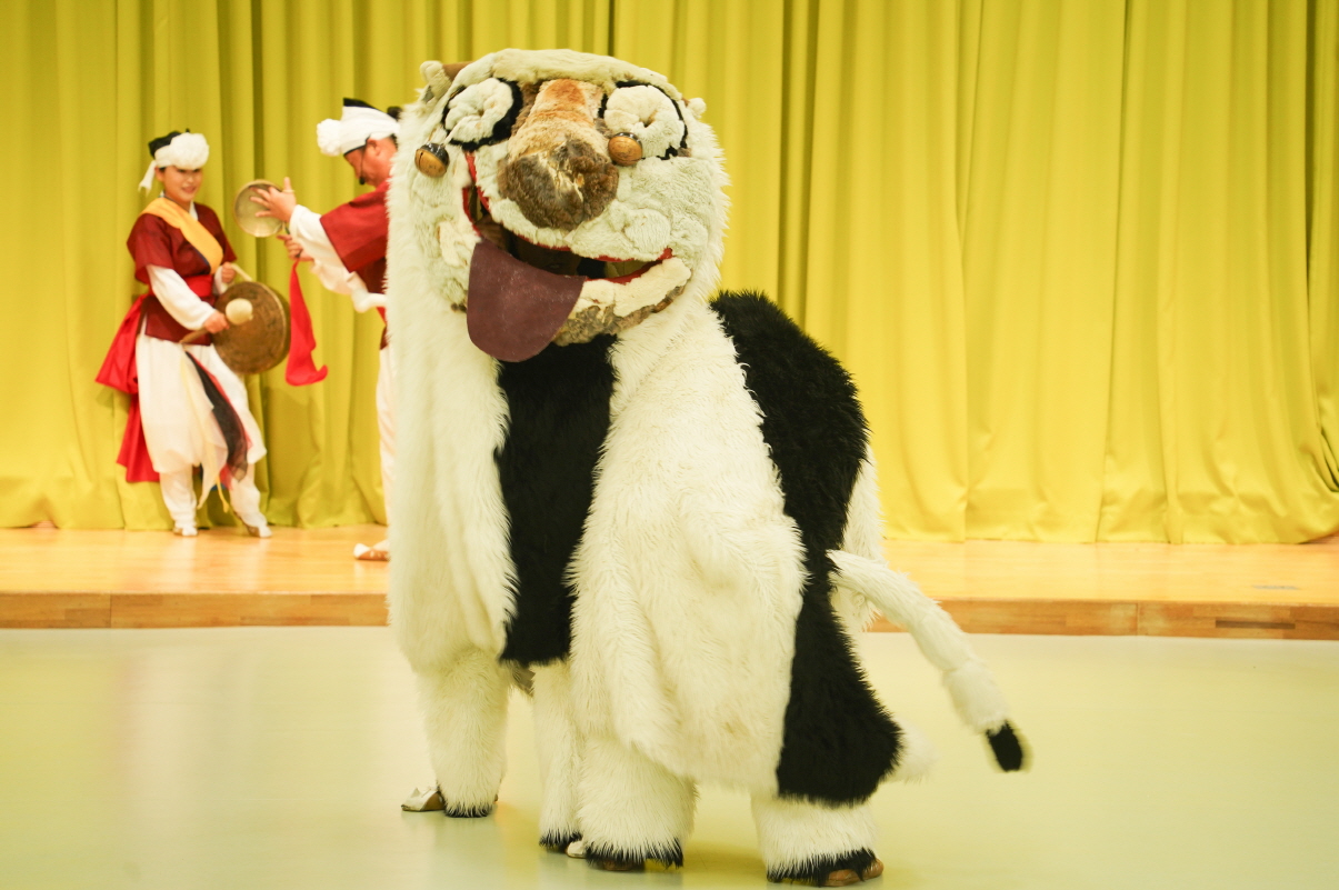 전주아름유치원에서 열린 유치원 공연 ‘풍물마당놀이 해피’ 중, 커다란 강아지 탈을 쓴 해피가 무대 중앙에서 관객을 향해 서 있고, 뒤편에서는 전통 의상을 입은 연희자들이 꽹과리와 징을 치며 공연을 펼치는 모습