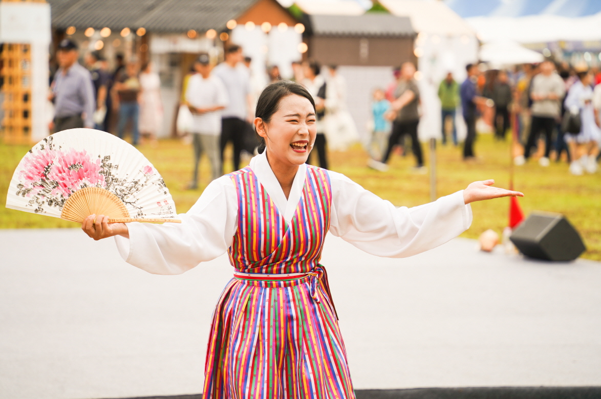 2025 안성 바우덕이 축제 마당놀이 복이팡팡 공연에서 부채를 든 소리꾼이 민요를 부르며 전통 한복을 입고 환하게 웃으며 관객과 소통하는 장면