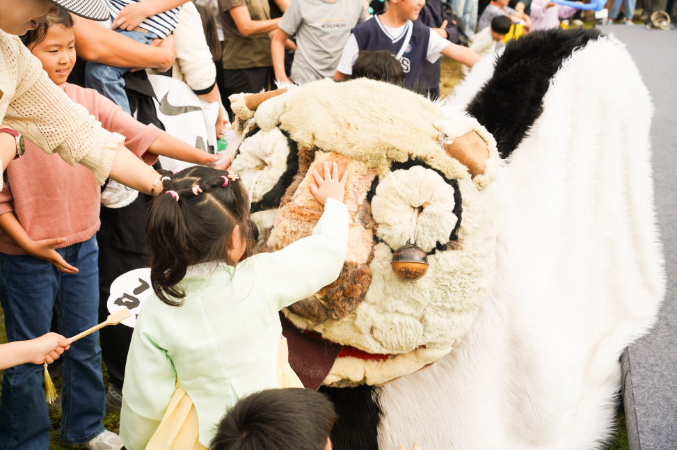 2025 안성 바우덕이 축제에서 아이가 풍물마당놀이 해피 공연의 강아지 탈 캐릭터를 만지며 즐거워하는 모습, 관객들과 아이들이 함께 교감하는 전통 공연 현장