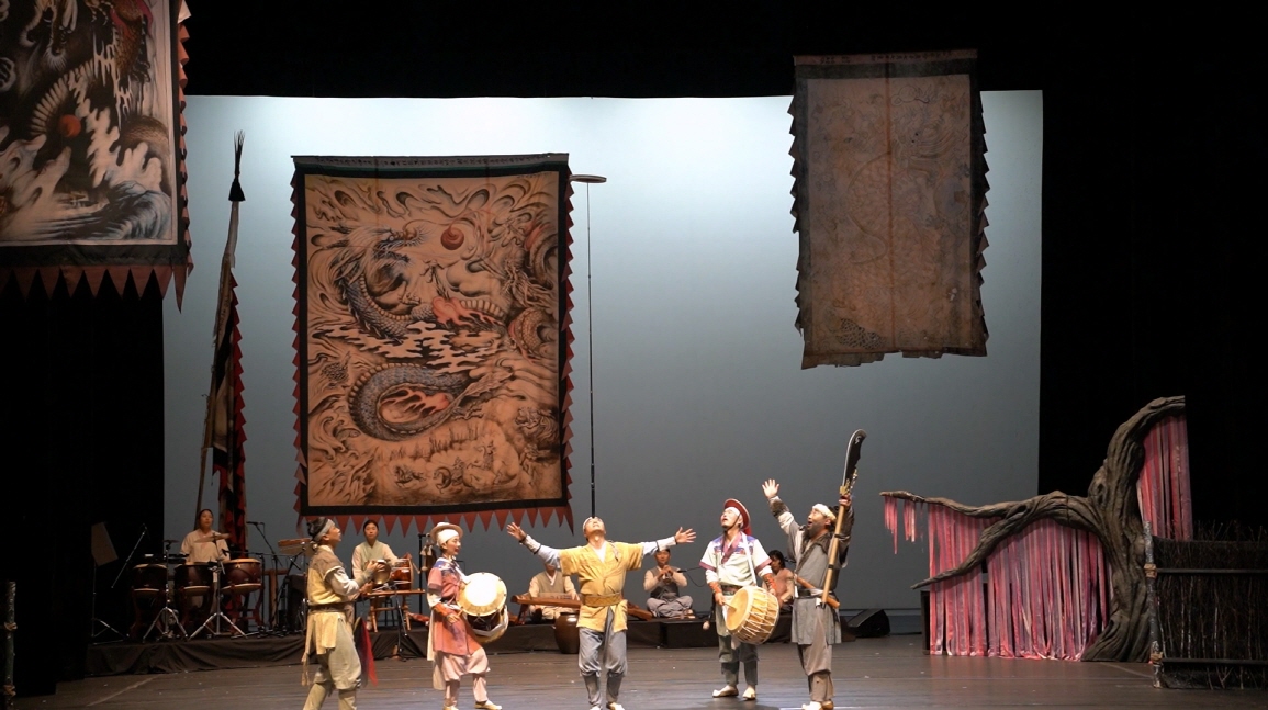 마당놀이 용을쫓는사냥꾼 공연에서 한 출연자가 머리 위 6m 높이의 막대에 버나를 올려 묘기를 부리며 손을 벌리고 있고, 사냥꾼들이 북과 징을 치며 무대를 고조시키는 장면