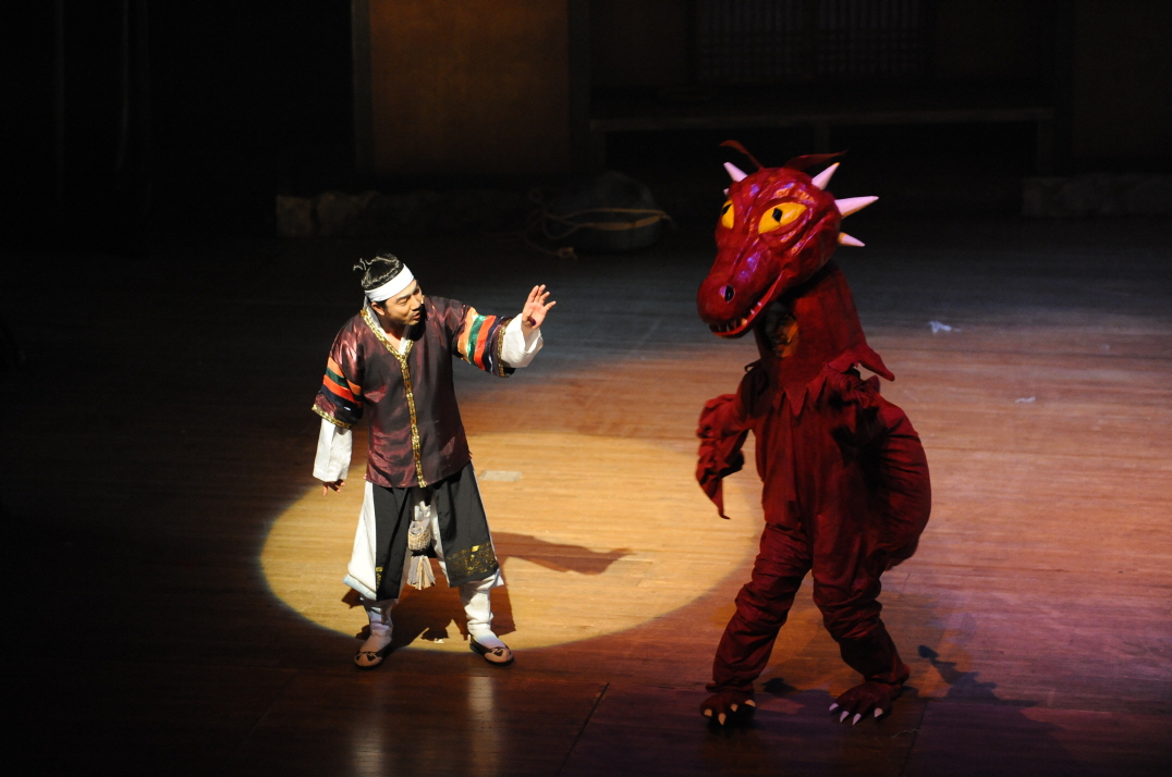 마당놀이 용을쫓는사냥꾼 공연에서 배우와 드래곤 캐릭터가 무대 위에서 서로 마주하며 코믹한 장면을 연출하는 모습