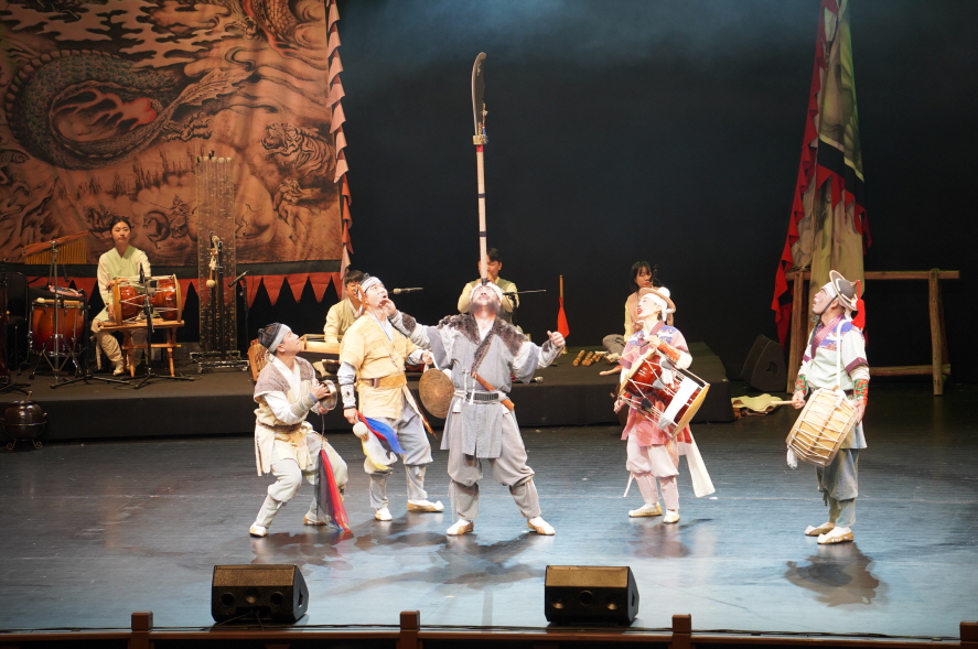 마당놀이 용을쫓는사냥꾼 공연에서 사냥꾼이 긴 무기를 턱으로 받쳐 올리며 전통기예를 선보이고, 연희자들이 북과 징을 치며 함께 장단을 맞추는 장면