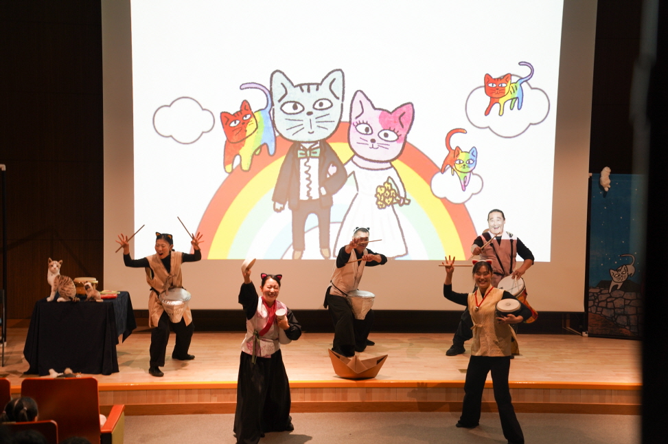 대구도서관 공연 어린이 마당놀이 ‘나는 고양이로소이다’ 공연 장면. 무대에서 배우들이 장단과 동작을 펼치며 연기하고 있고, 뒤 스크린에는 고양이 신랑·신부와 무지개, 여러 고양이 캐릭터가 등장하는 일러스트 애니메이션이 밝게 비춰지고 있다