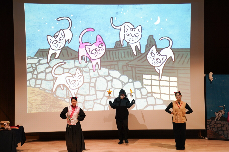 대구도서관 공연 어린이 마당놀이 ‘나는 고양이로소이다’ 공연 장면. 무대 앞에서 배우들이 고양이 캐릭터와 의상을 입고 연기하고 있으며, 뒤 스크린에는 여러 마리의 고양이 일러스트 애니메이션이 밝고 귀엽게 표현되어 있다.