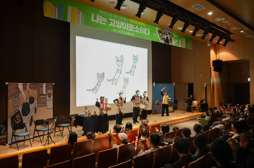 대구도서관 어린이 마당놀이 ‘나는 고양이로소이다’ 공연 장면. 무대에서 배우들이 북과 장단으로 공연을 펼치고 있으며, 스크린에는 고양이 일러스트 애니메이션이 재생되고 있다. 관객석에는 어린이와 가족들이 앉아 공연을 집중해서 관람하는 모습