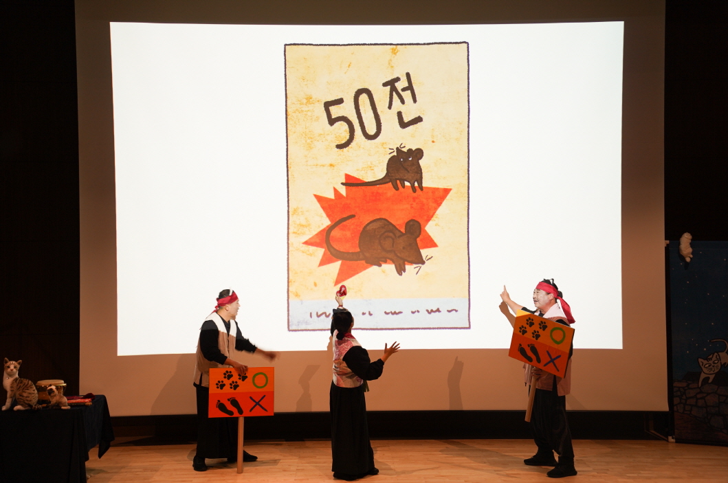 대구도서관 공연 어린이 마당놀이 ‘나는 고양이로소이다’ 공연 장면. 무대 중앙 스크린에 ‘50전’ 글씨와 쥐 그림이 나타난 장면이며, 배우들이 손팻말을 들고 등장인물 역할을 연기하고 있다