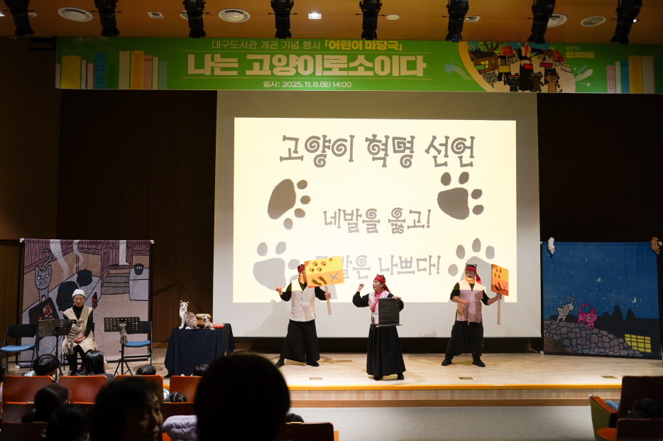 대구도서관 공연 어린이 마당놀이 ‘나는 고양이로소이다’ 공연 장면. 무대에서 배우들이 손팻말을 들고 고양이 혁명 선언 장면을 연기하고 있으며, 뒤 스크린에는 ‘고양이 혁명 선언, 네 발을 들고! 밥은 나쁘다!’라는 큰 글씨와 발바닥 그림이 비춰지고 있다.