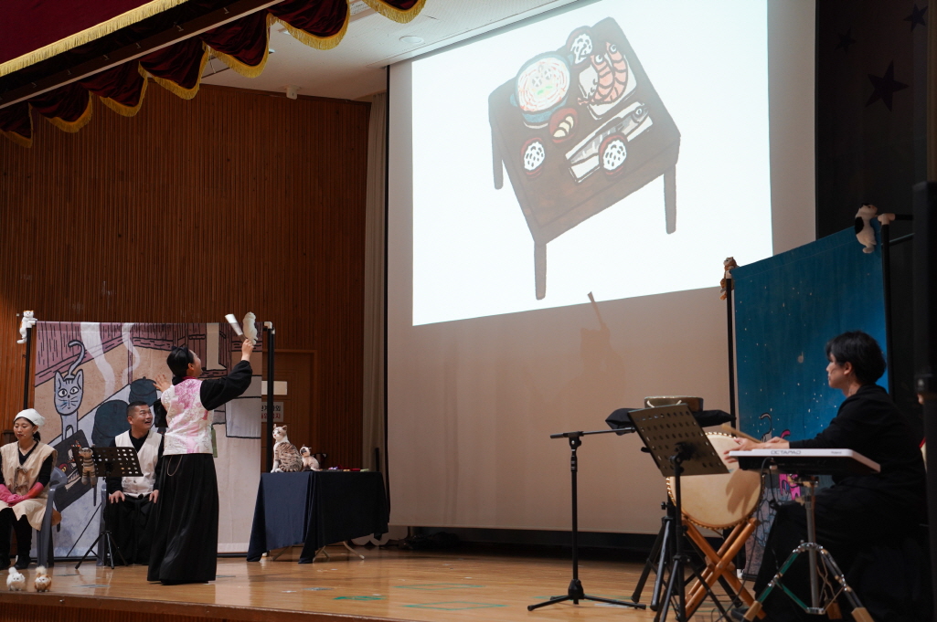 어린이 마당놀이 나는 고양이로소이다 공연에서 배우가 고양이 인형을 들고 연기하며, 무대 오른쪽에서는 라이브 반주자가 연주하고 있고 프로젝션 화면에 음식 일러스트가 비춰지는 장면