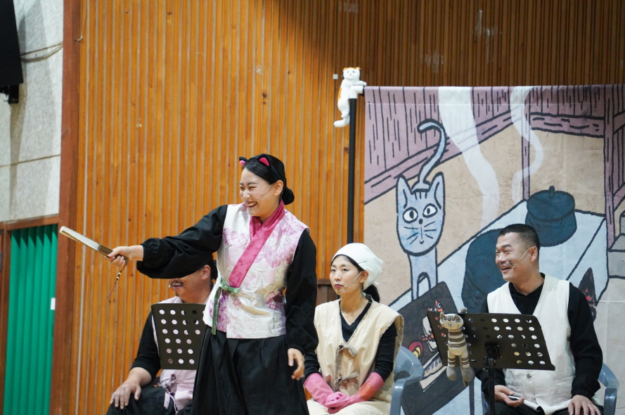 어린이 마당놀이 나는 고양이로소이다 공연에서 배우가 고양이 캐릭터로 무대 위에서 선창하며 관객과 소통하는 장면 – 일러스트 배경과 연주자들이 함께 공연 중