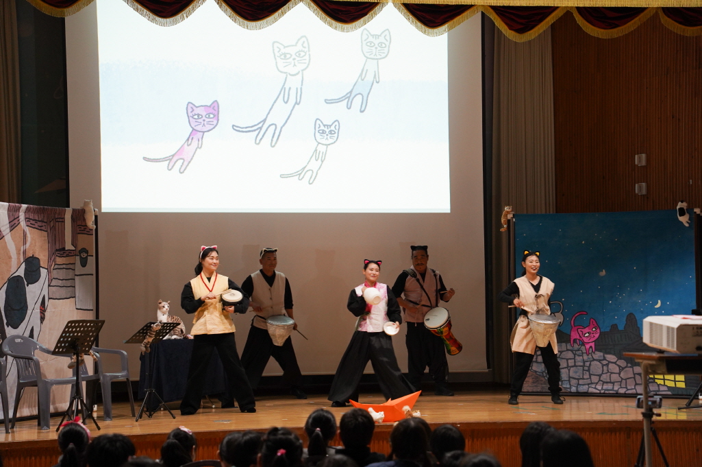 어린이 마당놀이 나는 고양이로소이다 공연에서 배우들이 고양이 귀를 달고 북과 소고를 연주하며 춤 장면을 펼치는 모습 — 무대 뒤 스크린에는 고양이 일러스트 애니메이션이 상영되고 객석의 학생들이 집중해 관람하는 장면