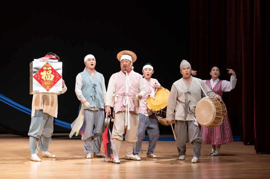 어르신 마당놀이 ‘복이 팡팡’에서 연희패가 북·장구·꽹과리를 치며 노래를 부르며 앞으로 걸어 나오는 행진 장면, 복 상자를 든 연희자가 함께 등장하는 한국 전통 공연