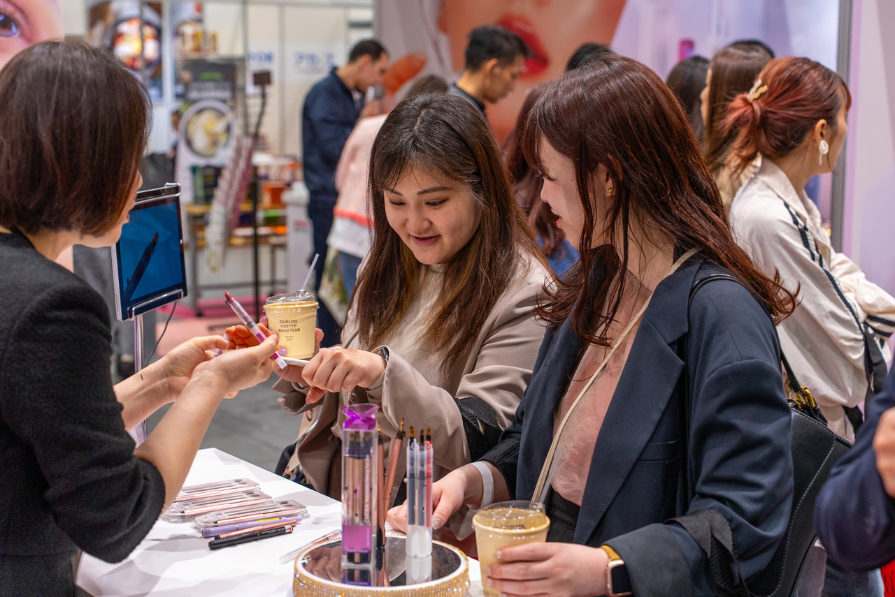 Visitors trying Korean cosmetics at a beauty booth, testing products with help from staff.