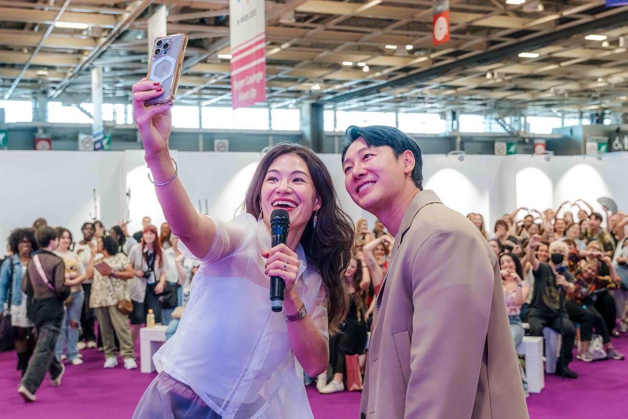 Visite surprise d'une célébrité coréenne lors de la Korea Expo 2025. Une fan prend un selfie avec un acteur coréen sur scène, montrant l'ambiance électrique