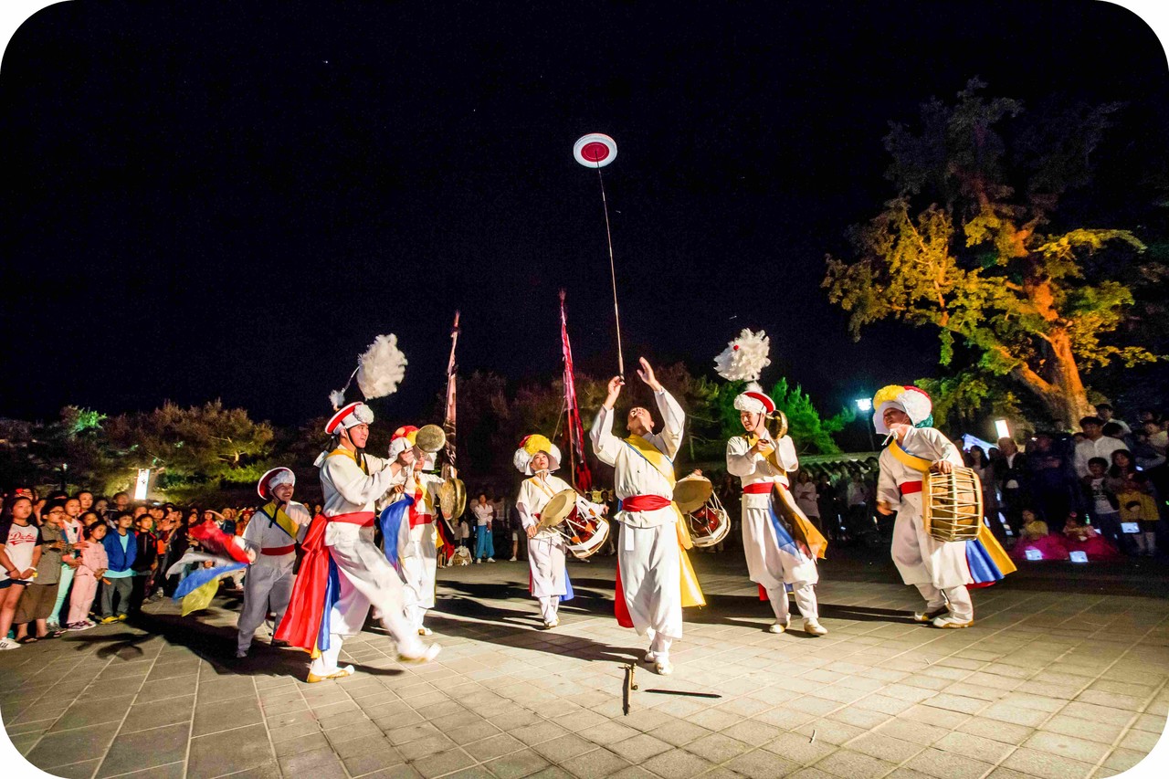 전주한옥마을 야간 축제 무대에서 연희자가 버나놀이 기예를 선보이며 남녀노소 관객들과 눈을 맞추고 즐겁게 소통하는 관객 참여형 공연 장면