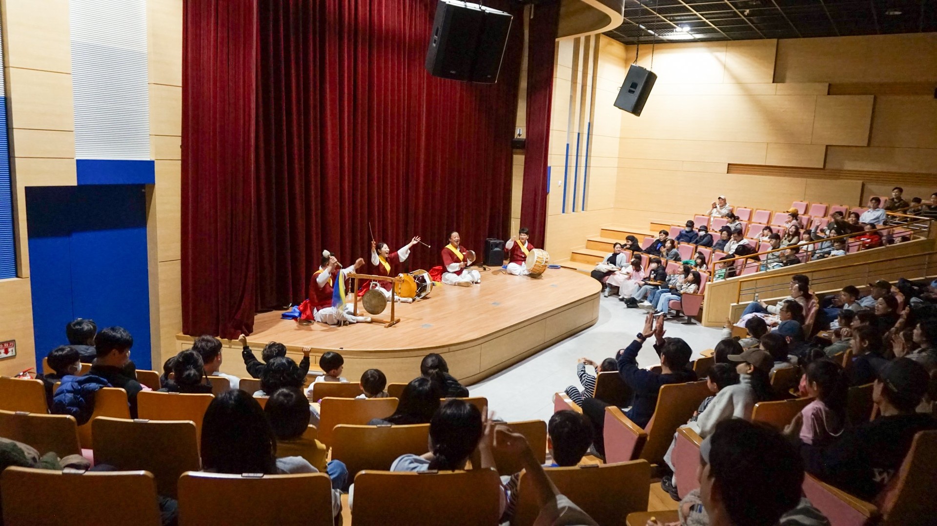 무대 위에서 괭과리, 장구, 북, 징을 연주하는 사물놀이 공연 현장과 경청하며 박수를 준비하는 100여 명의 관객들 모습