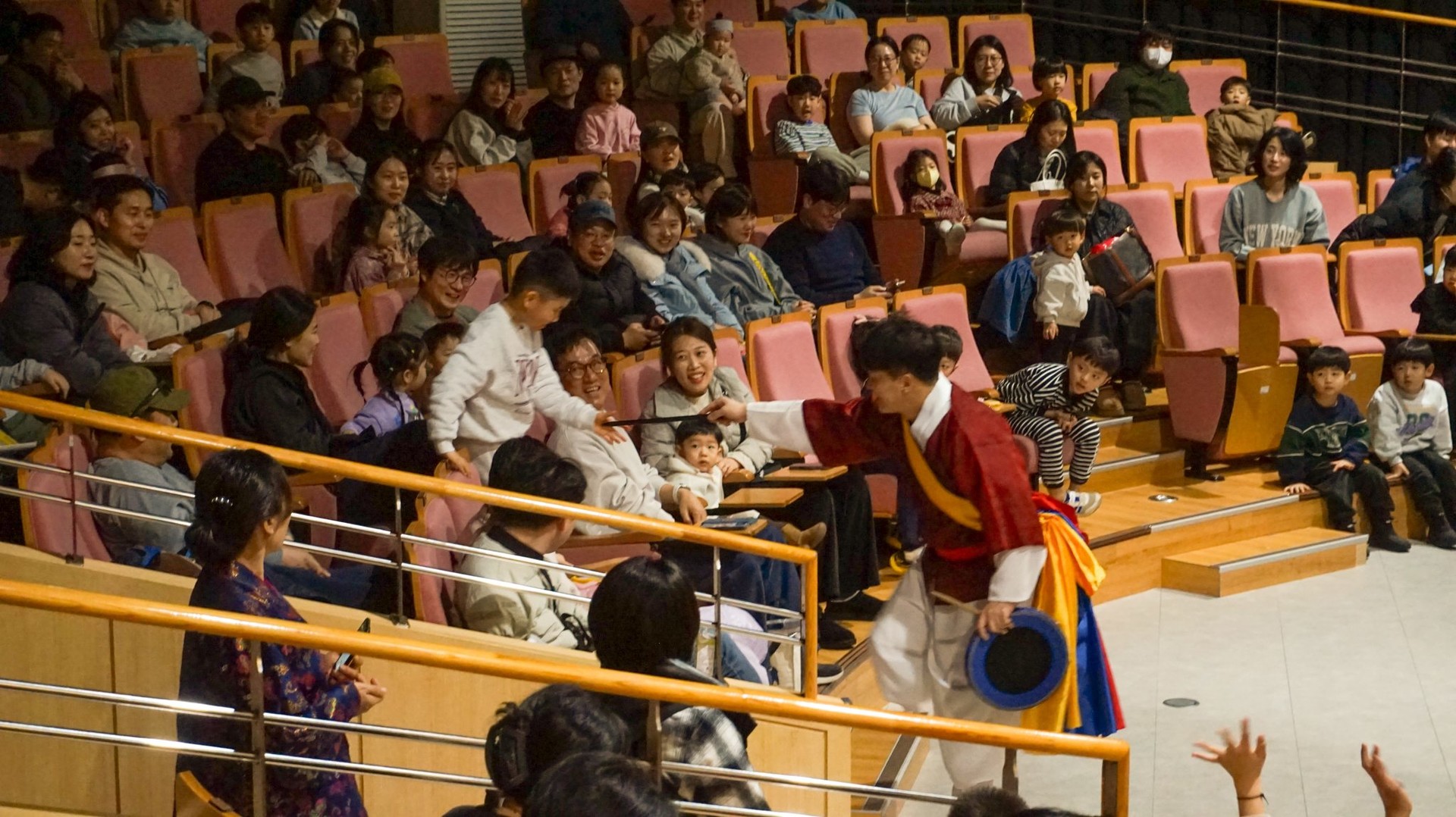 합굿마을 연주자가 객석의 어린이에게 북채를 건네며 직접 북을 쳐볼 수 있게 유도하는 모습과, 이 특별한 체험의 순간을 집중해서 지켜보는 관객들로 가득한 국립광주과학관 공연 현장