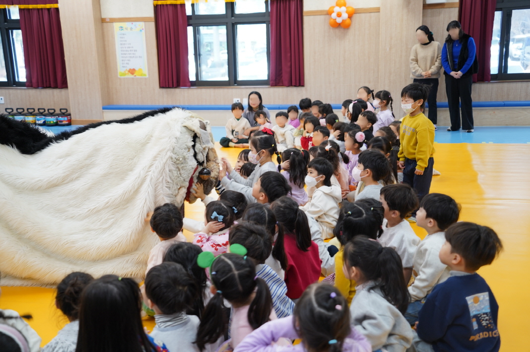 전주 푸른샘 유치원 강당에서 사회적기업 합굿마을의 강아지탈 캐릭터 '해피'가 아이들에게 가까이 다가가 인사하는 모습. 복실복실한 흰색 털의 해피와 이를 호기심 가득한 눈빛으로 바라보며 집중하고 있는 아이들의 생생한 공연 현장