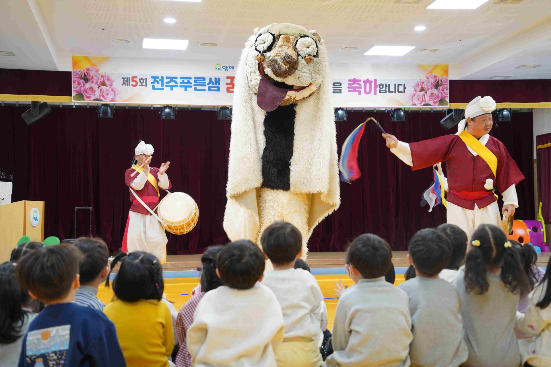 전주 푸른샘 유치원 무대 위에서 펼쳐지는 합굿마을 연희자들의 풍물놀이 공연. 북을 치는 연희자와 상모를 돌리는 출연자들 사이로 커다란 강아지탈 해피가 등장하여 아이들의 박수를 유도하며 흥겨운 분위기를 연출하는 모습