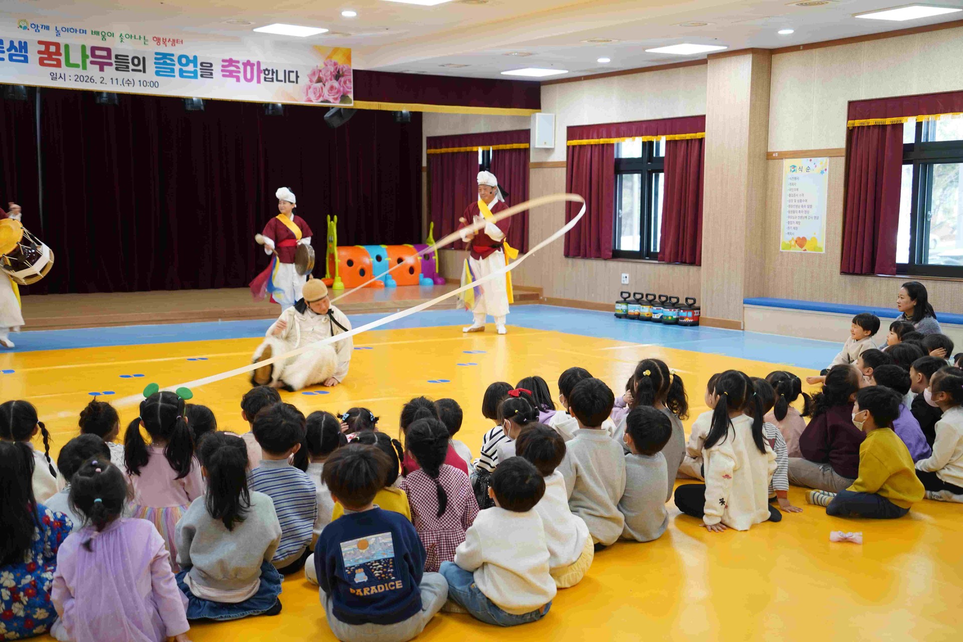 합굿마을 연희자가 무대 바닥에 앉아 긴 리본이 달린 상모를 돌리는 '열두발 놀이' 공연 장면. 화려한 궤적을 그리며 돌아가는 긴 리본과 이를 신기한 듯 바라보며 환호하는 전주 푸른샘 유치원 아이들의 뒷모습