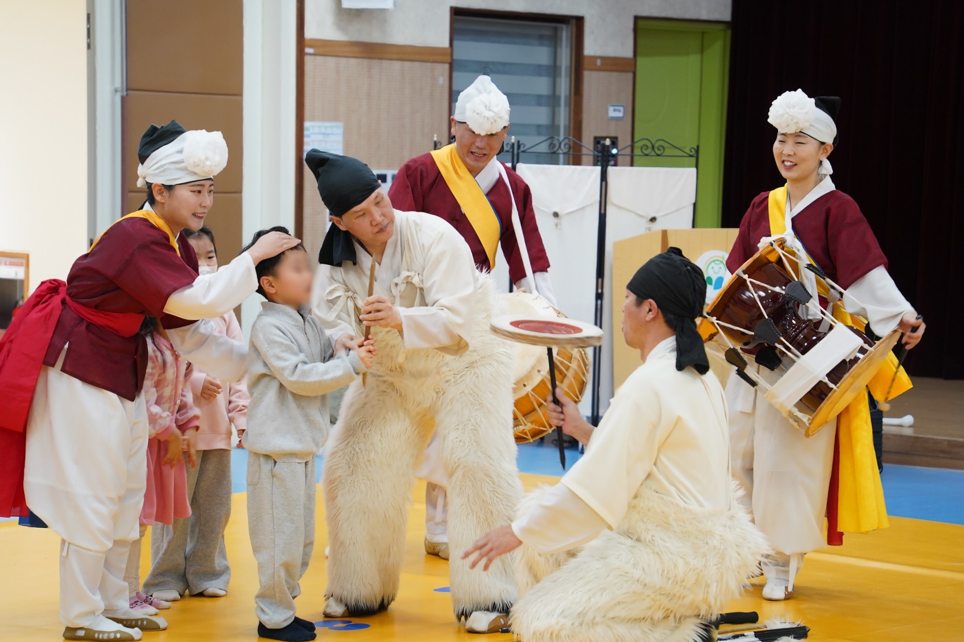 전주 푸른샘 유치원 버나놀이 체험 중, 출연자가 아이의 머리를 부드럽게 쓰다듬으며 격려하는 따뜻한 교감의 순간. 다른 연희자들이 주변에서 장구를 치며 응원하는 가운데, 아이가 직접 막대 위에서 접시(버나)를 돌려보며 전통 놀이의 재미를 느끼고 있다