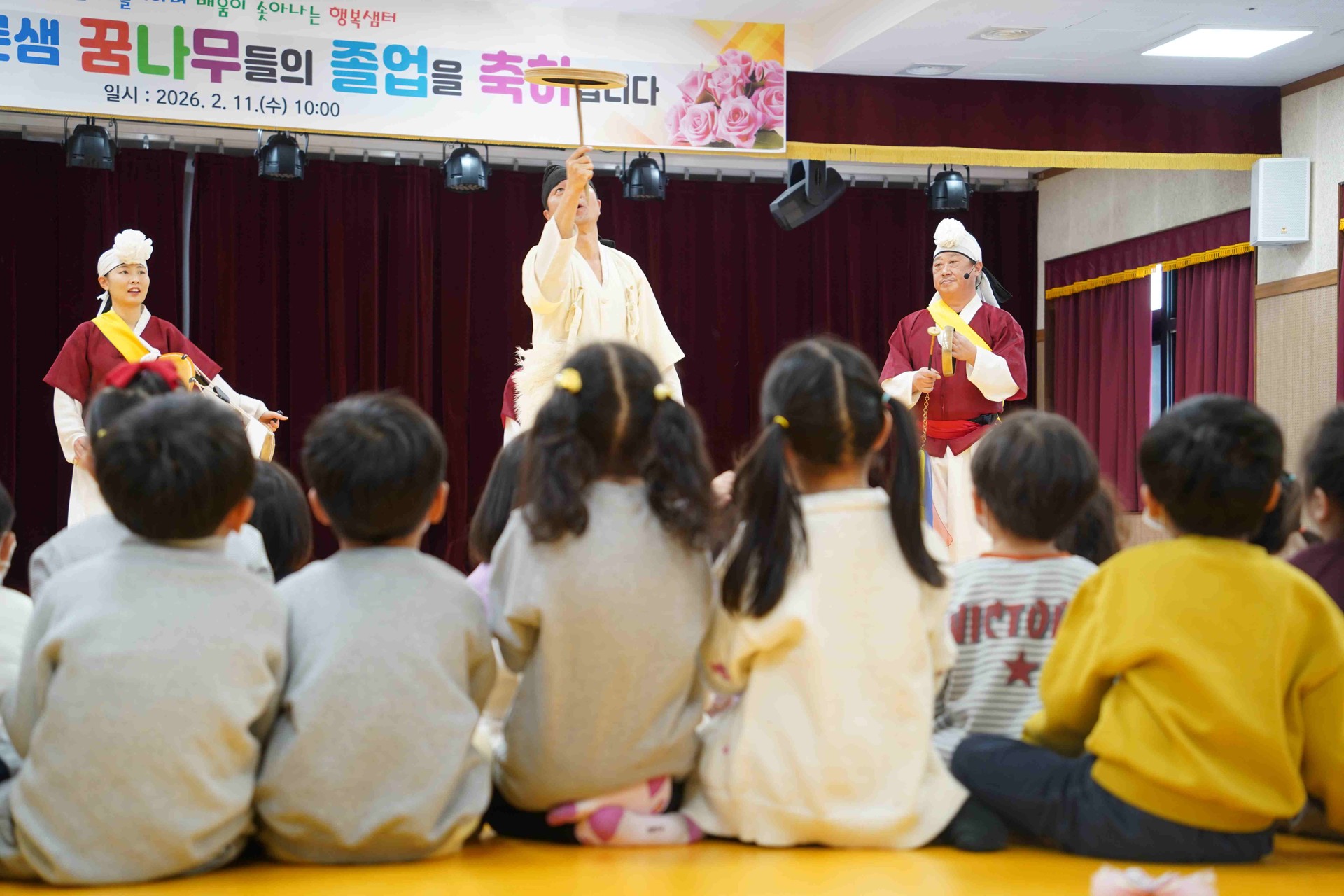 전주 푸른샘 유치원 공연 중, 연희자가 가느다란 막대 위에서 접시를 돌리는 '버나놀이' 기술을 선보이는 모습. 긴장감 넘치는 전통 묘기에 집중하며 무대 위 연희자의 손끝을 일제히 바라보고 있는 아이들의 뒷모습