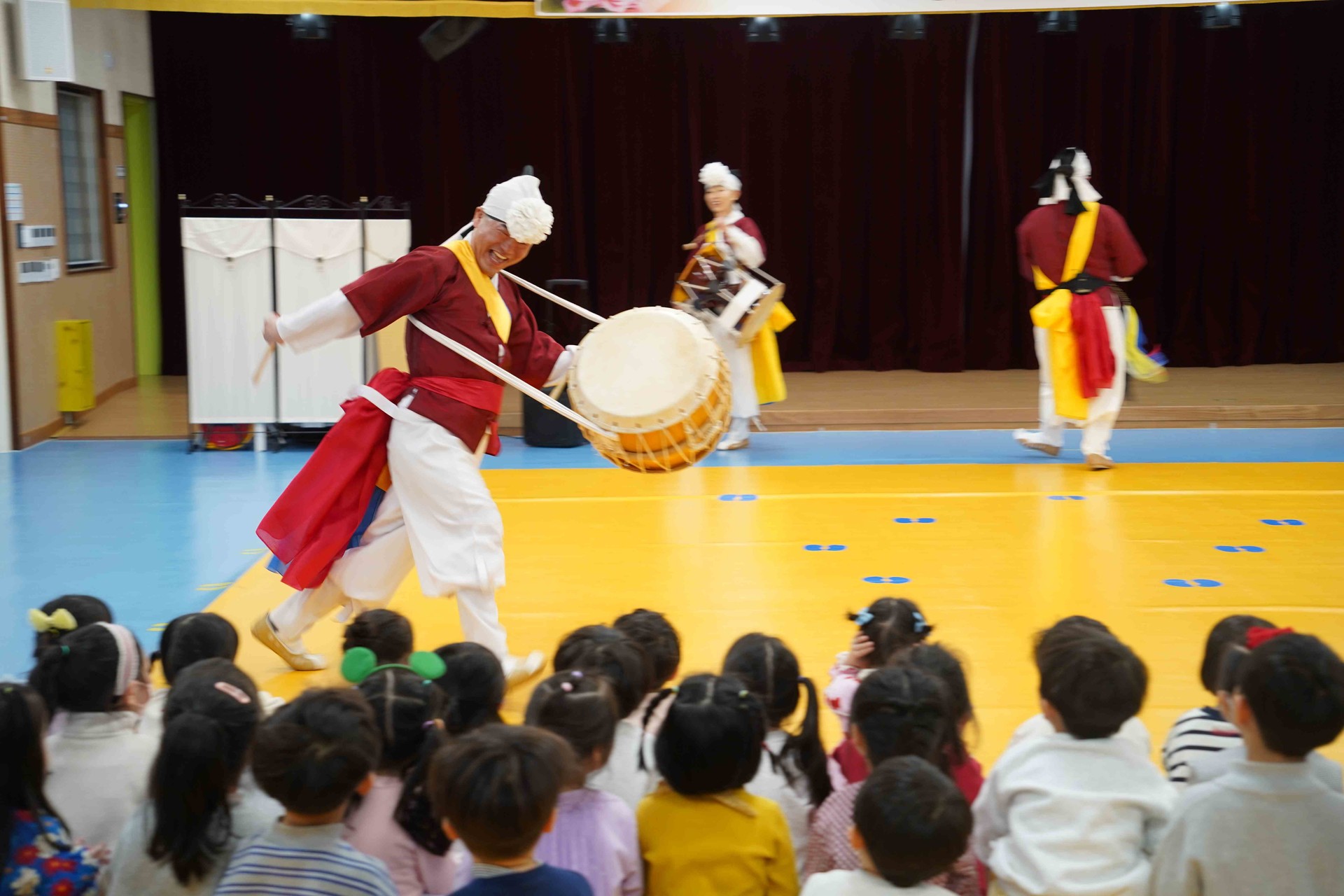 합굿마을 전문 연희자가 전주 푸른샘 유치원 아이들 앞에서 신명나게 북을 치며 뛰어다니는 역동적인 풍물놀이 장면. 수준 높은 예술성과 현장감 넘치는 연주에 매료되어 무대를 바라보는 아이들의 뒷모습