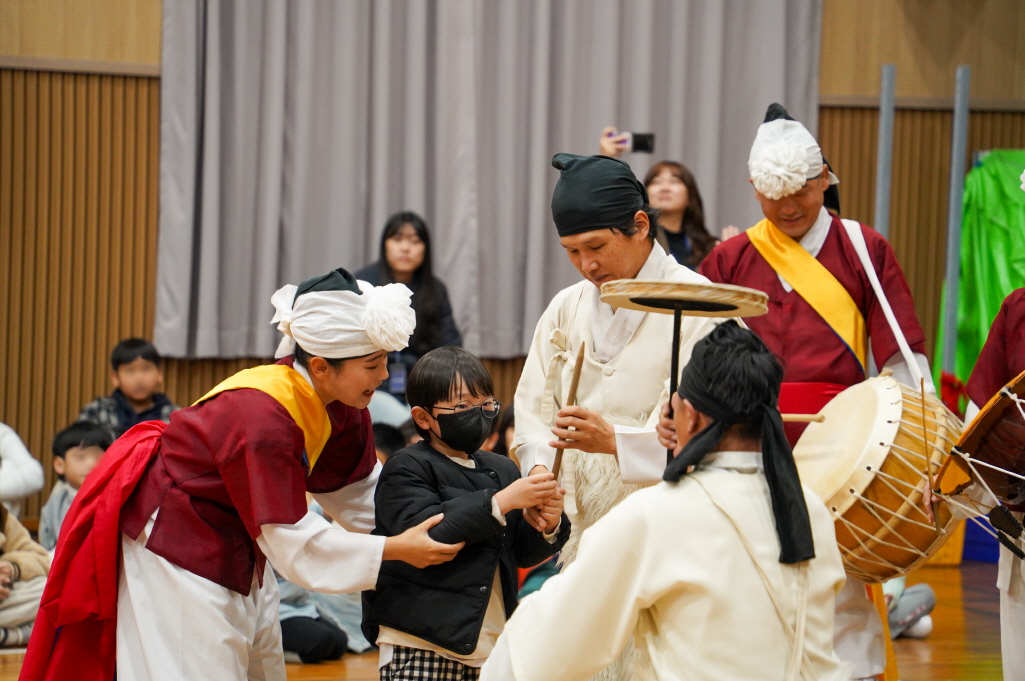 광주 비아초 병설유치원 강당에서 합굿마을 단원들이 어린이 관객에게 버나 막대기를 쥐여주며 직접 돌려볼 수 있게 도와주고, 아이가 집중해서 버나 체험에 참여하는 모습