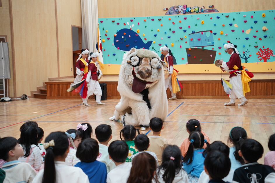 합굿마을 풍물마당놀이 사자탈춤 공연을 관람하는 목포부주초병설유치원 아이들. 고래 그림 클라이밍 벽 앞에서 해피 캐릭터가 익살스러운 표정으로 춤추는 모습